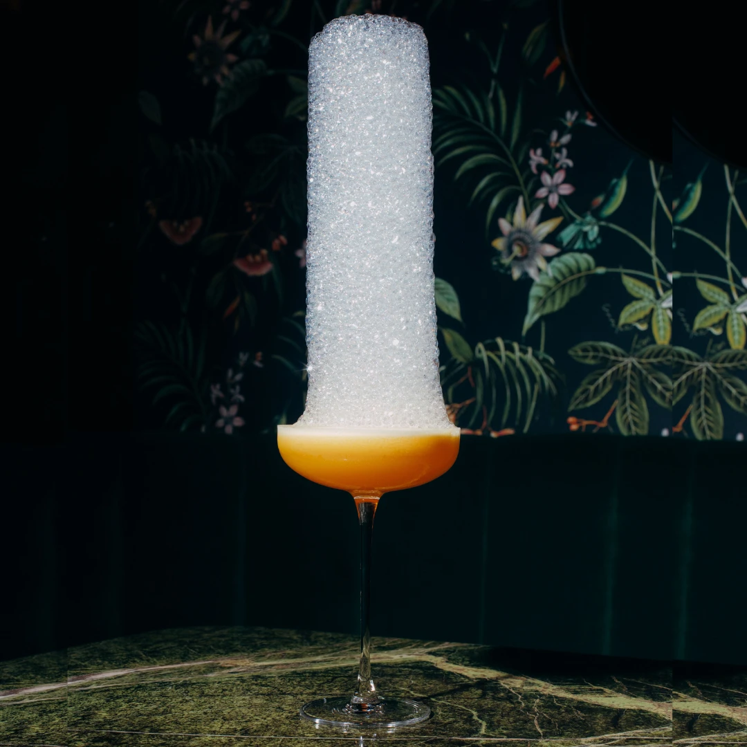 Cocktail mit hohem Schaumtopping auf Marmortisch vor floraler Wand.